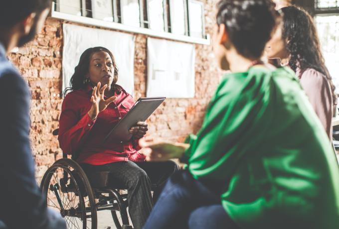 A woman leads a meeting in a brick-walled room while three attendees look on.